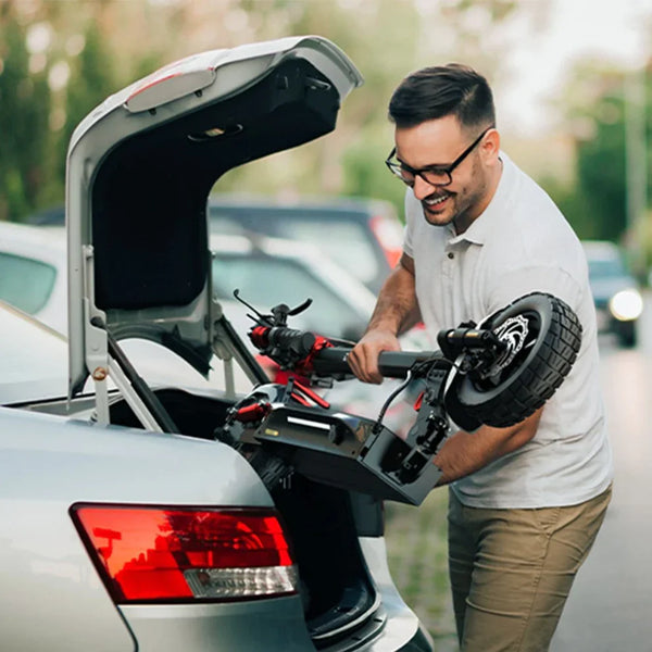 Na sliki je električni skiro, model Engwe Y600S. Vidimo moškega, ki ga nalaga v prtljažnik avtomobila. Skrbno ga drži in se smeška, kar nakazuje, da je zadovoljen z uporabo skiroja. V ozadju so drugi avtomobili, kar kaže na urbano okolje.