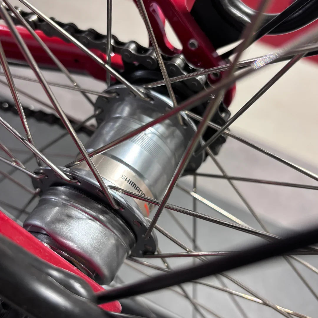 Izdelek je električno kolo, znamke Fischer, model Cita 1.0. Na sliki je prikazan del zadnjega kolesa, kjer je vidna os, natančneje Shimano hub, ter del verige in platišča. Kolo je v rdeči barvi, kar daje dinamičen videz.