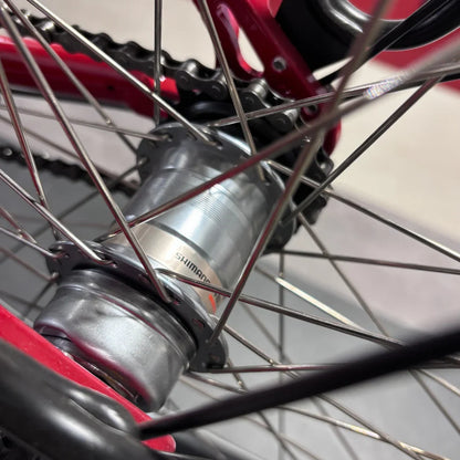 Izdelek je električno kolo, znamke Fischer, model Cita 1.0. Na sliki je prikazan del zadnjega kolesa, kjer je vidna os, natančneje Shimano hub, ter del verige in platišča. Kolo je v rdeči barvi, kar daje dinamičen videz.