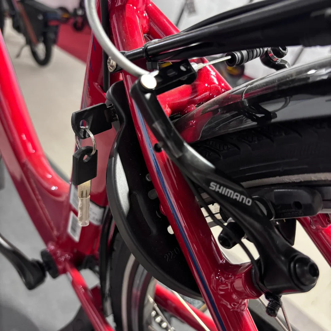 Izdelek na sliki je električno kolo znamke Fischer, model Cita 1.0. Na sliki je vidna rdeča okvirna konstrukcija kolesa, del zavornega sistema, ki je označen z znamko Shimano, ter ključ, ki je pritrjen na okvir. Kolo je zasnovano za udobno vožnjo in vključuje elemente, ki zagotavljajo varnost in zanesljivost.