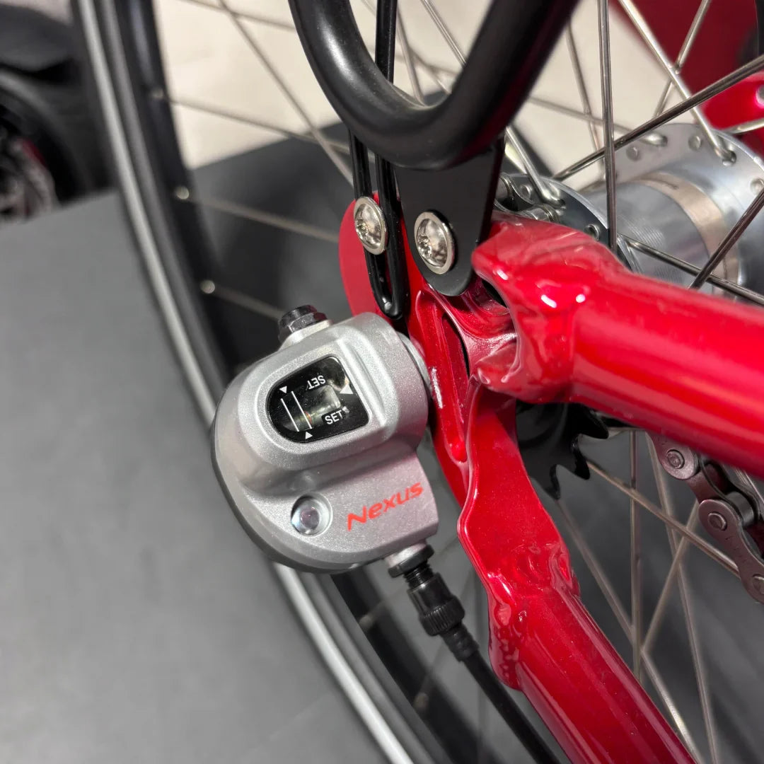 Izdelek na sliki je električno kolo, znamke Fischer, model Cita 1.0. Na sliki je prikazan del kolesa, kjer je vidna prestavna naprava Nexus, ki omogoča enostavno prestavljanje. Rdeča barva okvirja in detajli na mehanizmu dodajajo estetski videz kolesu.