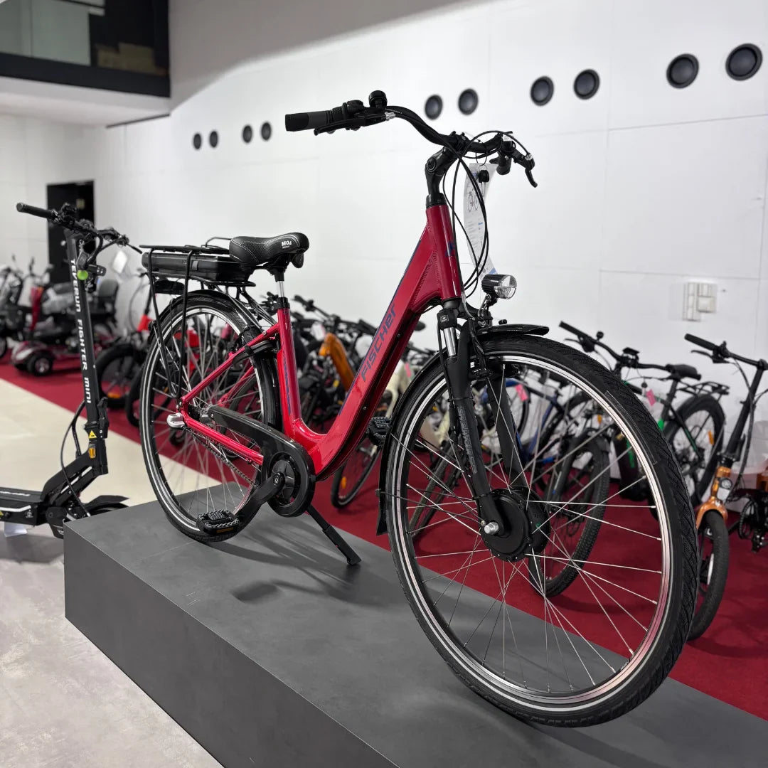 Na sliki je električno kolo znamke Fischer, model Cita 1.0. Kolo je v svetlo rdeči barvi in ima udoben sedež ter dodatno opremo, kot je nosilec za prtljago. V ozadju so vidni tudi drugi modeli koles in električnih skirojev, kar nakazuje, da gre za prodajno mesto.
