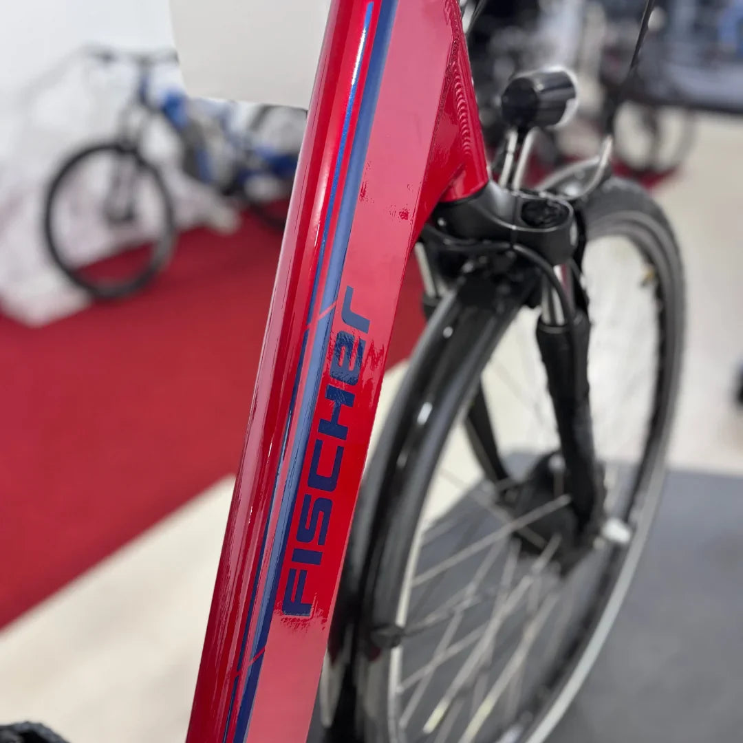 Izdelek na sliki je električno kolo, model Fischer Cita 1.0. Na sliki je vidna rdeča okvir kolesa z značilnim logotipom znamke Fischer, ki se nahaja na okvirju. V ozadju so vidni še drugi kolesi, kar kaže, da je kolo postavljeno v prodajnem prostoru.