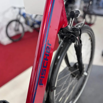 Izdelek na sliki je električno kolo, model Fischer Cita 1.0. Na sliki je vidna rdeča okvir kolesa z značilnim logotipom znamke Fischer, ki se nahaja na okvirju. V ozadju so vidni še drugi kolesi, kar kaže, da je kolo postavljeno v prodajnem prostoru.