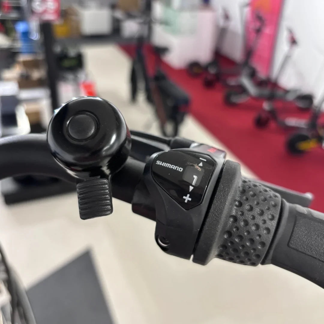 Izdelek je električno kolo znamke Fischer, model Cita 1.0. Na sliki je prikazan del krmila, kjer je vidna ročica za prestavljanje Shimano in zvonec. V ozadju se nahajajo drugi električni skiroji, kar nakazuje, da je slika posneta v trgovini s kolesi in električnimi vozili.