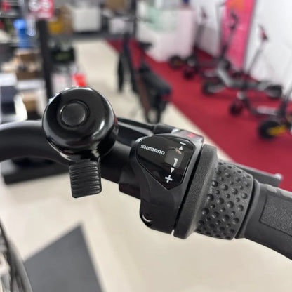 Izdelek je električno kolo znamke Fischer, model Cita 1.0. Na sliki je prikazan del krmila, kjer je vidna ročica za prestavljanje Shimano in zvonec. V ozadju se nahajajo drugi električni skiroji, kar nakazuje, da je slika posneta v trgovini s kolesi in električnimi vozili.