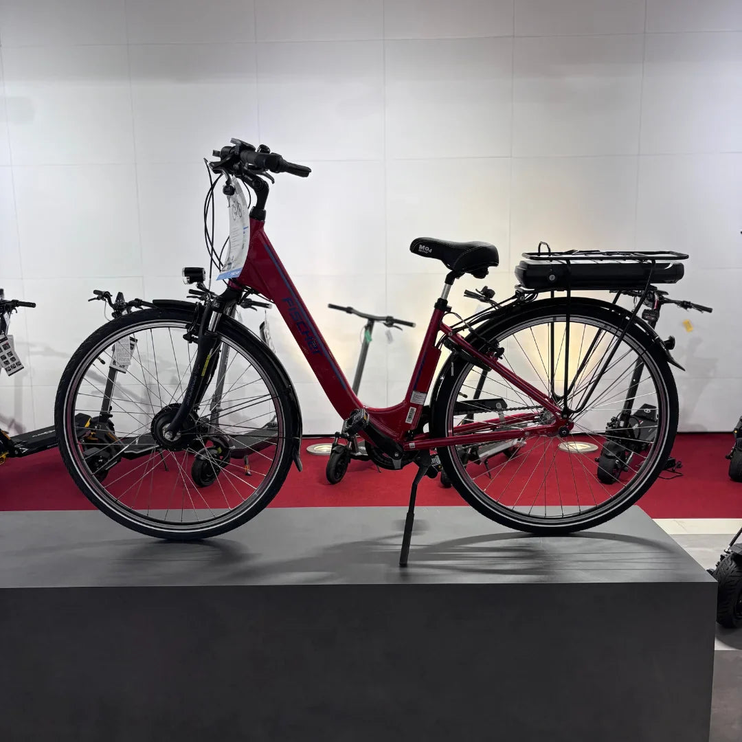 Na sliki je električno kolo znamke Fischer, model Cita 1.0. Kolo je rdeče barve in ima udoben okvir z nizkim prečnim članom, kar olajša vstopanje. Na zadnjem delu kolesa je nameščena baterija, kar nakazuje na njegovo električno delovanje. V ozadju so vidni tudi drugi električni skiroji, kar daje občutek o raznolikosti električnih prevoznih sredstev.