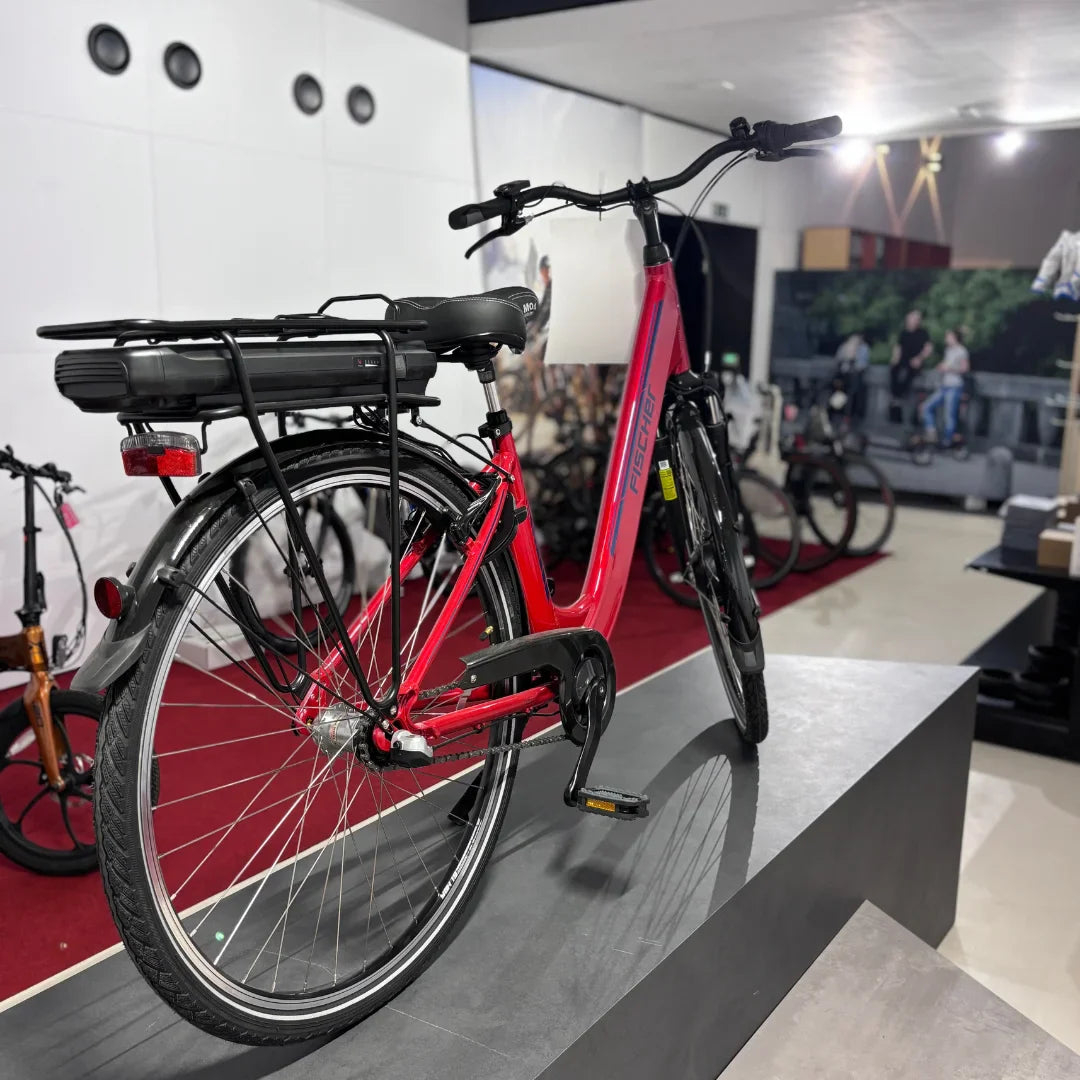 Izdelek na sliki je električno kolo znamke Fischer, model Cita 1.0. Kolo je v svetlo rdeči barvi in ima nameščen akumulator na zadnjem delu okvirja. Vidna je tudi klasična zasnova z dvema kolesoma, krmilom in pedalom, kar je značilno za električna kolesa. V ozadju so vidni drugi kolesi in prostoren prostor za razstavo.