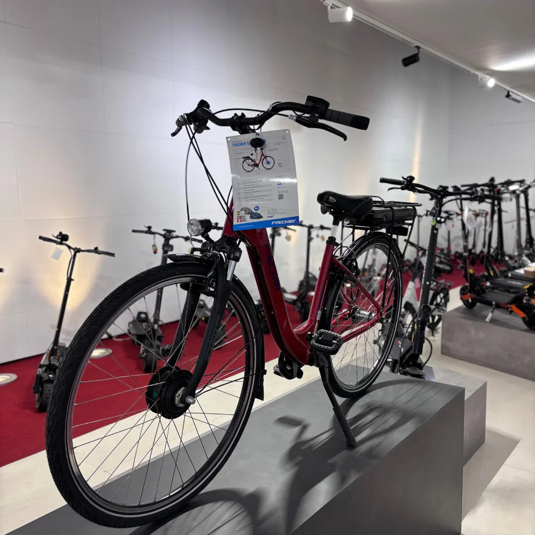 Izdelek na sliki je električno kolo, znamke Fischer, model Cita 1.0. Na sliki je vidno kolo v rdeči barvi, ki je postavljeno na stojalo, ob njem pa je informativna oznaka. V ozadju so vidni tudi drugi električni skiroji. Kolo ima klasično obliko okvirja in je opremljeno z električnim motorjem ter baterijo na zadnjem delu.