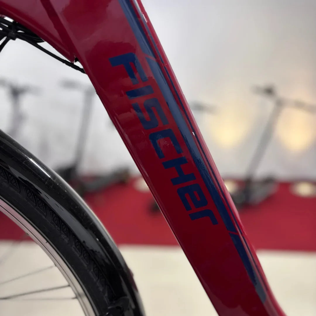 Na sliki je električno kolo znamke Fischer, model Cita 1.0. Vidna je rdeča barva okvirja s plavo oznako znamke Fischer. V ozadju so delno vidni tudi električni skiroji, kar nakazuje, da je slika posneta v trgovini ali na razstavi.