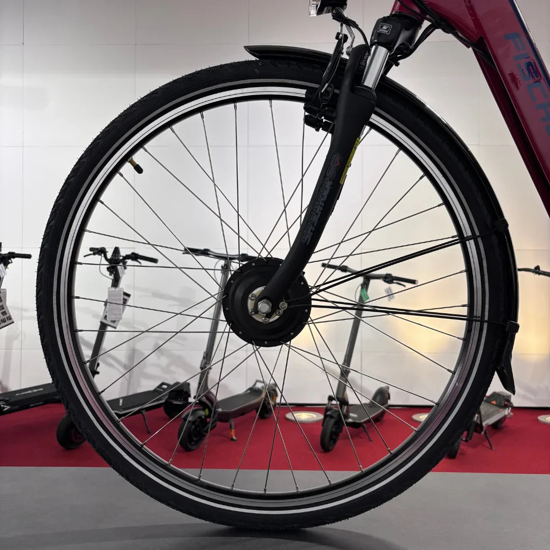 Na sliki je električno kolo znamke Fischer, model Cita 1.0. Vidna je sprednja pnevmatika in vilice kolesa, kar nakazuje na robustno zasnovo, primerna za mestno vožnjo. V ozadju so vidni tudi električni skiroji, kar daje občutek sodobnega urbanega okolja.