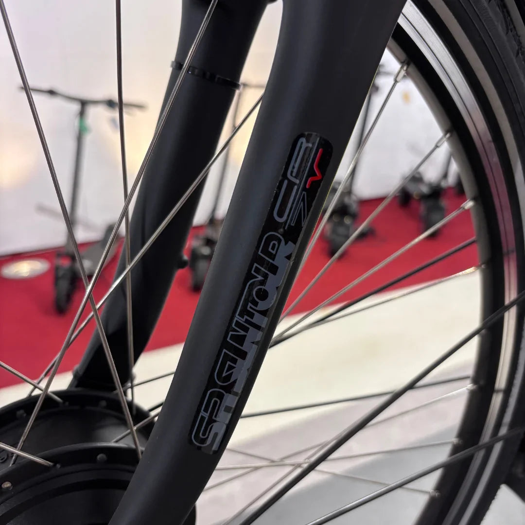 Izdelek na sliki je električno kolo, znamke Fischer, model Cita 1.0. Na sliki je prikazan del kolesa, natančneje vilice, ki so izdelane iz črnega materiala. Vidni so tudi deli kolesa, kot so žice in del motorja, v ozadju pa so vidni električni skiroji.