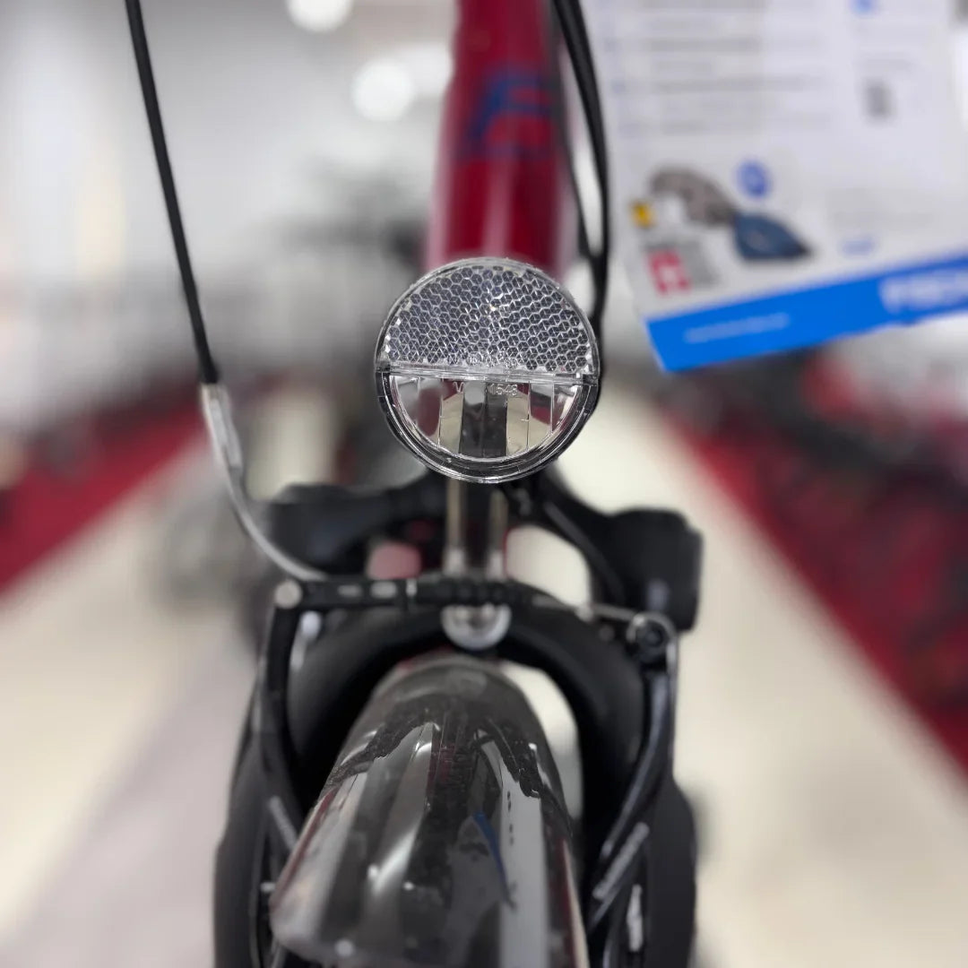 Na sliki je električno kolo, model Fischer Cita 1.0. Vidna je sprednja luč, ki je nameščena na kolesu, ter del okvirja in krmila. Kolo je v rdeči barvi, kar je lahko opazno na okvirju. Ozadje slike prikazuje druge kolesa v trgovini.