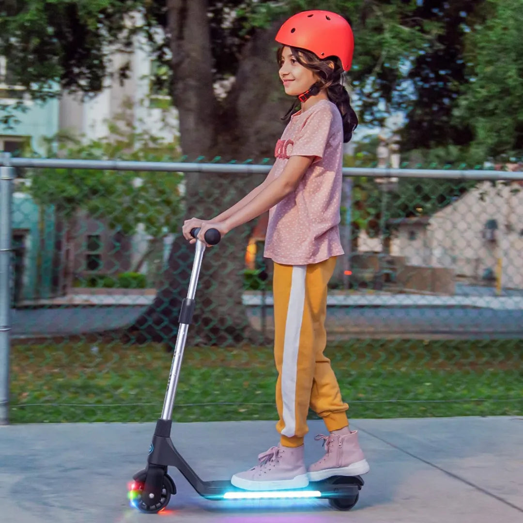 Na sliki je električni skiro znamke Gyroor, model H30. Vidimo deklico, ki vozi skiro po pločniku, oblečeno v udobna oblačila in zaščitno čelado rdeče barve. Scoot je opremljen z LED lučmi na sprednjem delu, kar dodaja vizualno privlačnost in povečuje varnost pri vožnji. V ozadju je park s drevesi in ograjo.