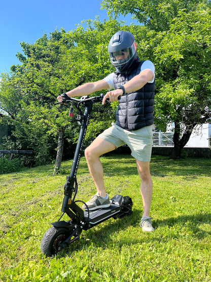 Na sliki je električni skiro model Teverun Fighter Mini Pro 2025. Prikazuje moškega, ki stoji na skiroju na travnati površini, oblečenega v kratke hlače in majico, ter nosi čelado. V ozadju so drevesa in modro nebo, kar nakazuje lepo vreme. Električni skiro ima robustno konstrukcijo in široke pnevmatike, kar je značilno za tovrstne naprave.