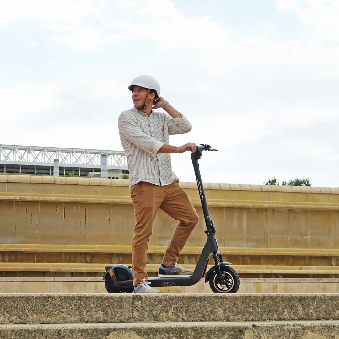 Na sliki je električni skiro znamke Joyor, model Litego. Vidimo moškega, ki stoji na skiroju in nosi čelado, kar nakazuje na varno vožnjo. Okolica je mestna, z arhitekturnimi elementi v ozadju, kar daje občutek prostornosti in svobode pri vožnji.