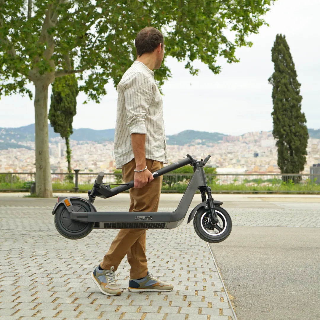 Na sliki je električni skiro, model Joyor Litego. Oseba ga nosi, kar kaže na njegovo prenosljivost in lahkost. V ozadju je mestna pokrajina z drevesi in hribi, kar ustvarja prijetno urbano okolje. Skratka, izdelek je zasnovan za enostavno mobilnost v mestnih območjih.