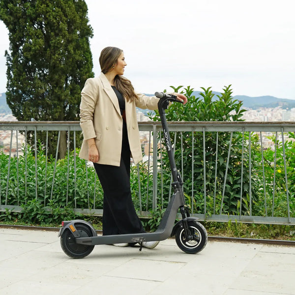 Na sliki je električni skiro znamke Joyor, model Litego. Predstavljen je v urbanem okolju, kjer ga uporablja ženska, oblečena v moderen plašč. Skrbi za eleganten videz, medtem ko se skiro opira ob ograjo, v ozadju pa je vidna mestna panorama in zelenje.