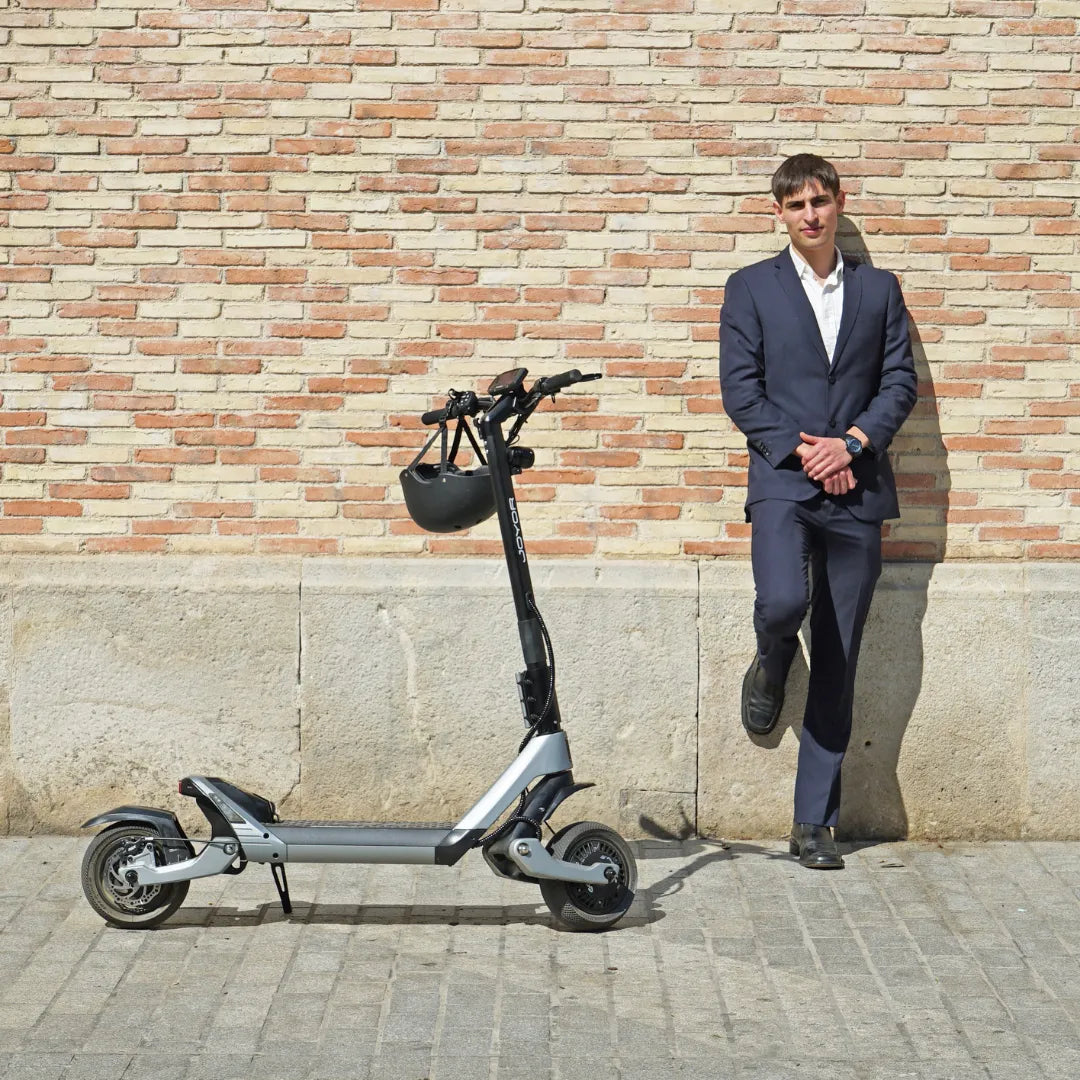 Moški v obleki se naslanja na steno poleg električnega skiroja Joyor Luxerider.