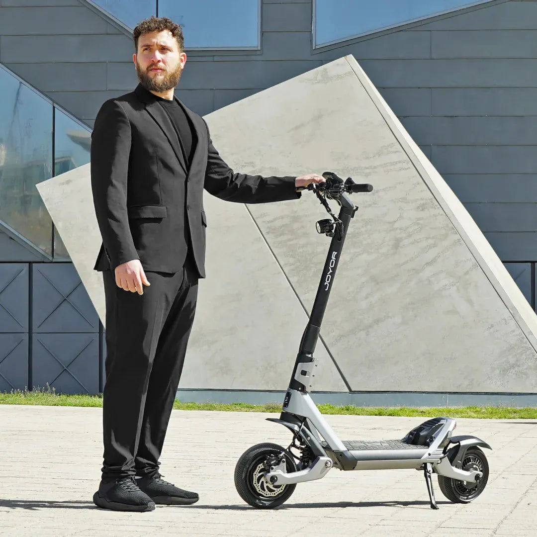 Moški v obleki stoji poleg električnega skiroja Joyor Luxerider pred moderno zgradbo.