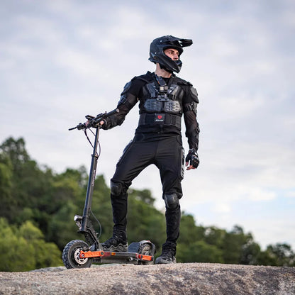 Na sliki je prikazan električni skiro znamke Joyor, model S-Pro. Oseba na skiroju nosi zaščitno opremo, vključno s čelado in zaščitnimi oblačili, kar kaže na namen vožnje v zahtevnejših pogojih. Scoot je postavljen na skalnato površino, v ozadju pa so vidne zelene drevesne krošnje, kar nakazuje na naravno okolje.