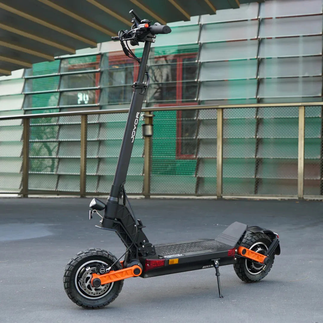 Izdelek na sliki je električni skiro znamke Joyor, model S-Pro. Na sliki je prikazan skiro s črnim okvirjem, širokimi pnevmatikami in svetlo oranžnimi detajli. Ima ergonomsko oblikovan krmilo in široko platformo za udobno vožnjo. Ozadje prikazuje moderno arhitekturo, kar daje sliki sodoben videz.