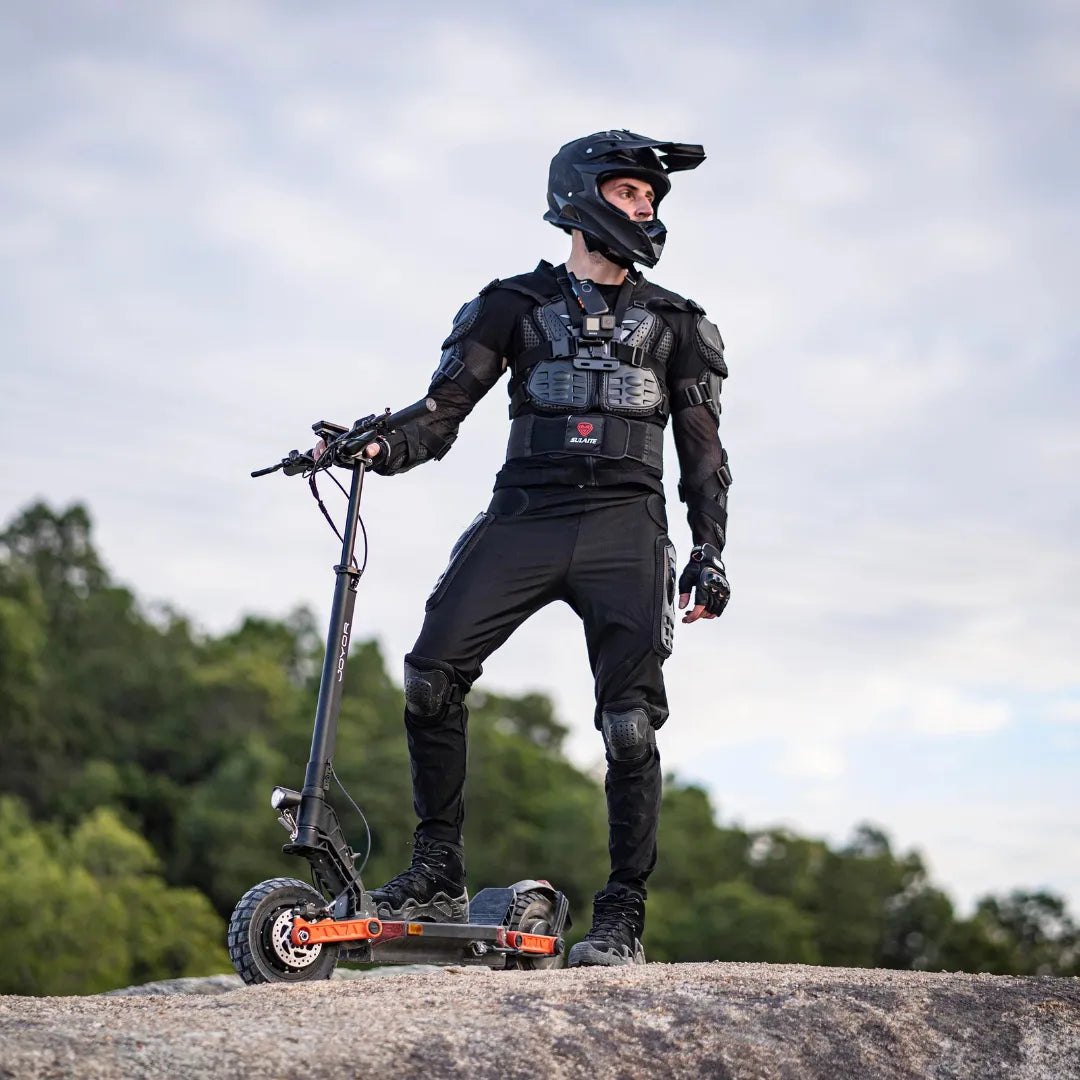 Na sliki je električni skiro, model Joyor S5 Z. Vidimo voznika, oblečenega v zaščitno opremo, vključno s čelado in ščitniki, ki stoji na skiroju na skalnati površini. Ozadje je naravno, z zelenjem, kar nakazuje na zunanjo okolico. Električni skiro ima robustno zasnovo, primeren za terensko vožnjo.