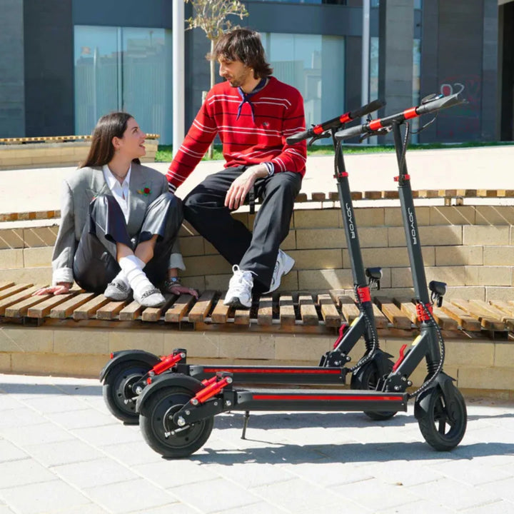 Ženska in moški sedita na klopi ob dveh električnih skirojih Joyor G5.