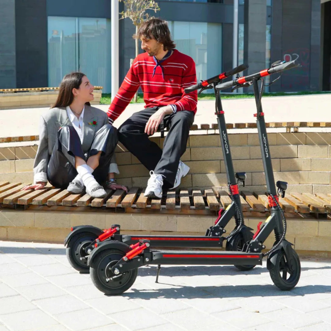 Na sliki je prikazan električni skiro znamke Joyor, model G5. Vidni sta dva električna skiroja, ki sta postavljena ob klopi, kjer sedita moški in ženska. Oboje je oblečeno v sproščena oblačila, kar ustvarja prijetno vzdušje. Skirava imata črno in rdečo barvno kombinacijo ter so zložena, kar kaže na njihovo prenosljivost.