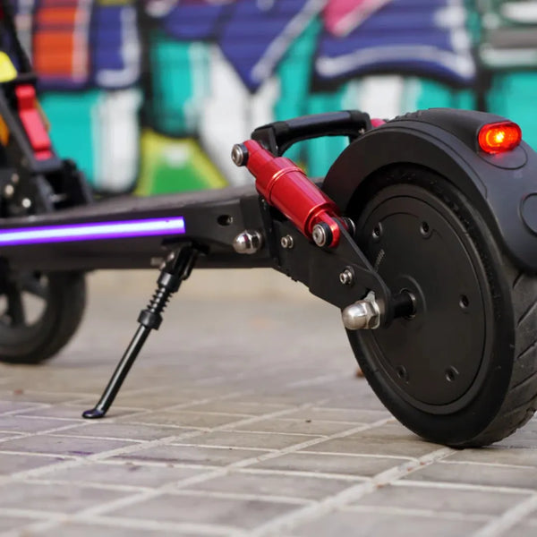 Izdelek je električni skiro znamke Joyor, model G5. Na sliki je vidna zadnja kolesna enota skiroja, ki vključuje motor, amortizer in LED lučko. Opremljen je s sistemom za blaženje udarcev ter ima robustno gumo, kar omogoča udobnejšo vožnjo. V ozadju je vidna grafitna stena, kar daje sliki urban videz.