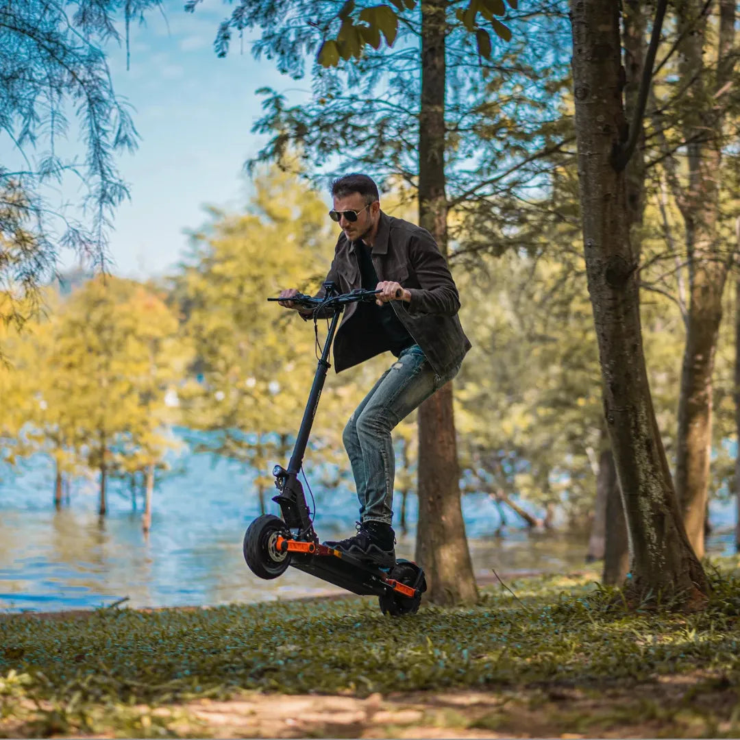 Na sliki je električni skiro, model Joyor S10 S-Z. Vidimo moškega, ki vozi skiro v naravnem okolju, ob vodi in med drevesi. Ima na sebi sončna očala in se zdi, da uživa v vožnji. Scoot je črn z oranžnimi detajli, kar mu daje športen videz.
