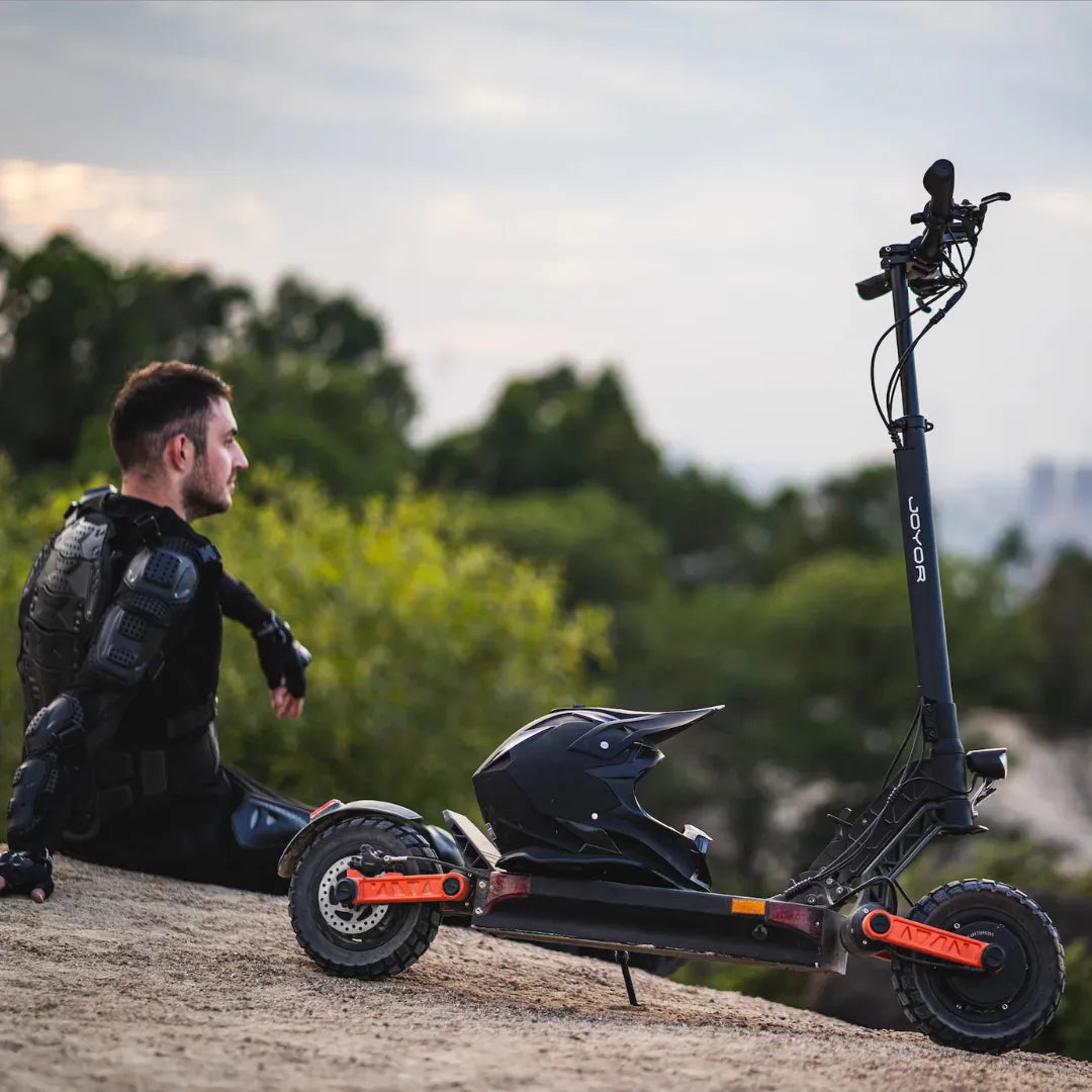 Oseba v zaščitni opremi sedi ob električnem skiroju Joyor S10 S Z na makadamski poti.