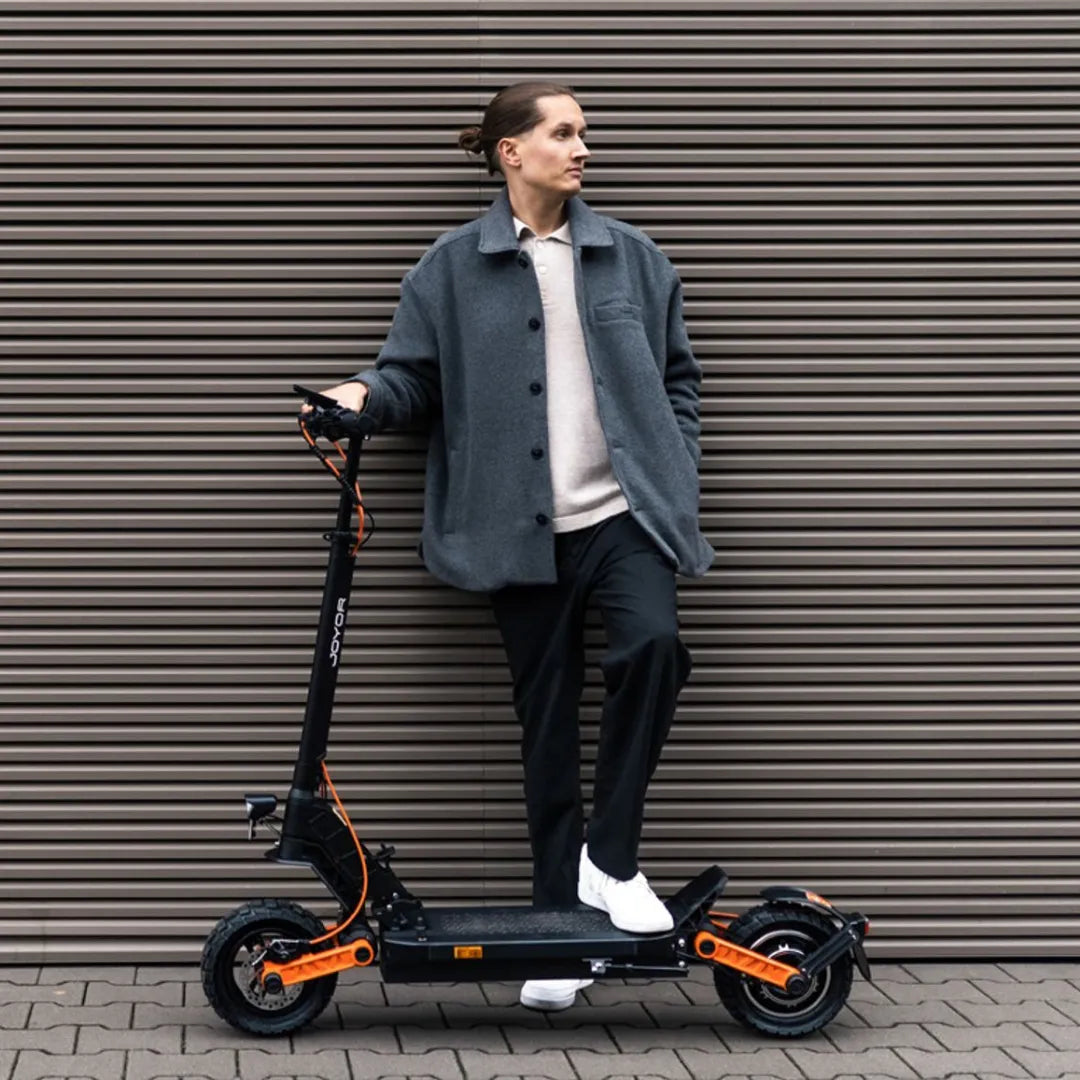 Oseba v sivi jakni stoji ob električnem skiroju Joyor S10 S Z ob teksturirani steni.