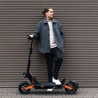 Oseba v sivi jakni stoji ob električnem skiroju Joyor S10 S Z ob teksturirani steni.