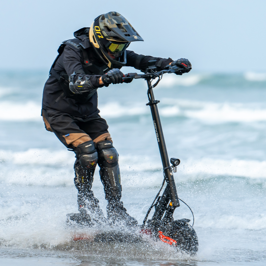 The product in the picture is an electric scooter, model Joyor T10. The picture shows a person riding the scooter along the coast, surrounded by water and sea waves. The person is wearing protective gear, including a helmet and knee pads, which indicates active and dynamic use of the scooter in demanding conditions.