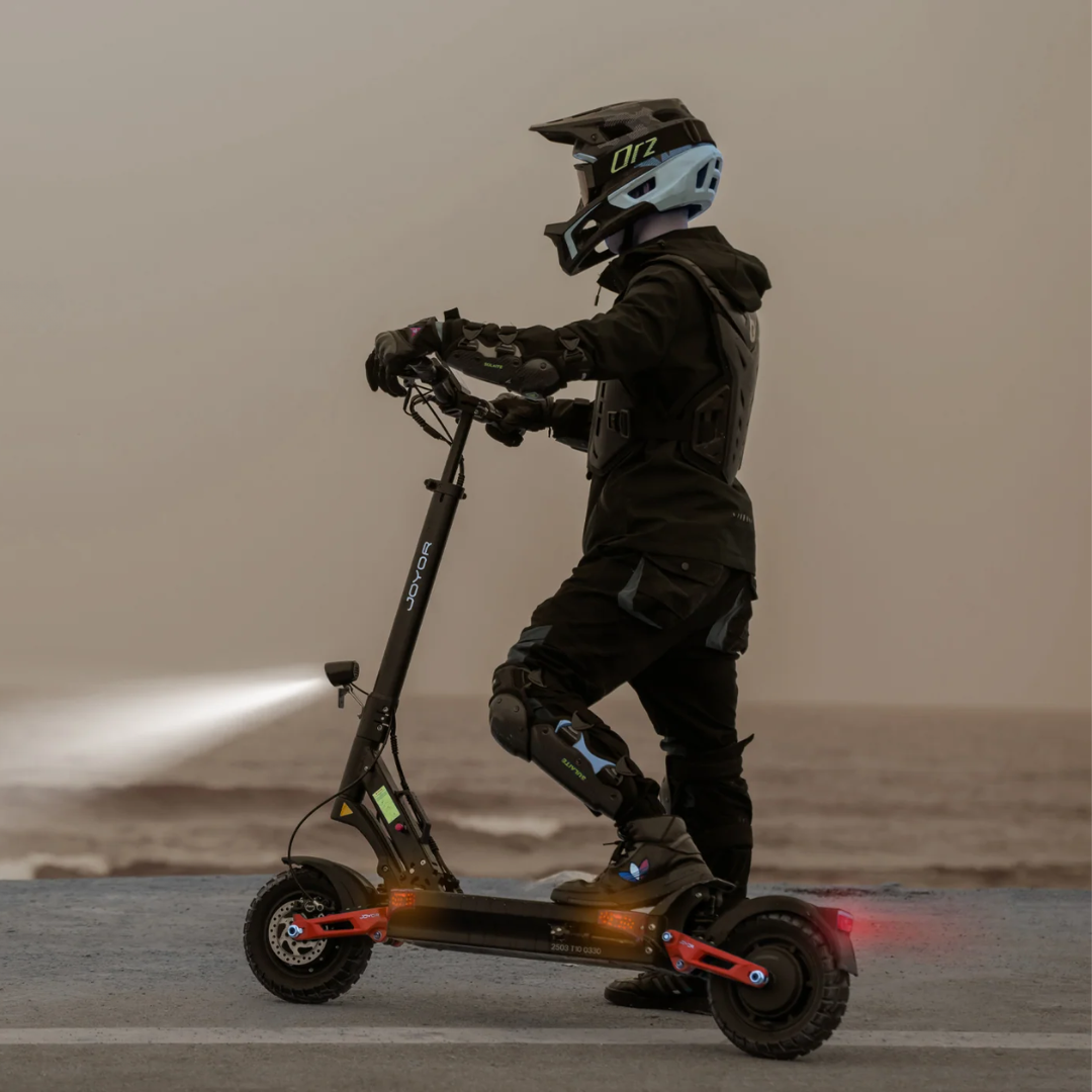The image shows a Joyor electric scooter, model T10. The user is standing on the scooter, wearing protective gear, including a helmet and protective pads. In the background, the coast and sea are visible, and the scooter has a built-in light, suggesting it can be ridden in low-light conditions.