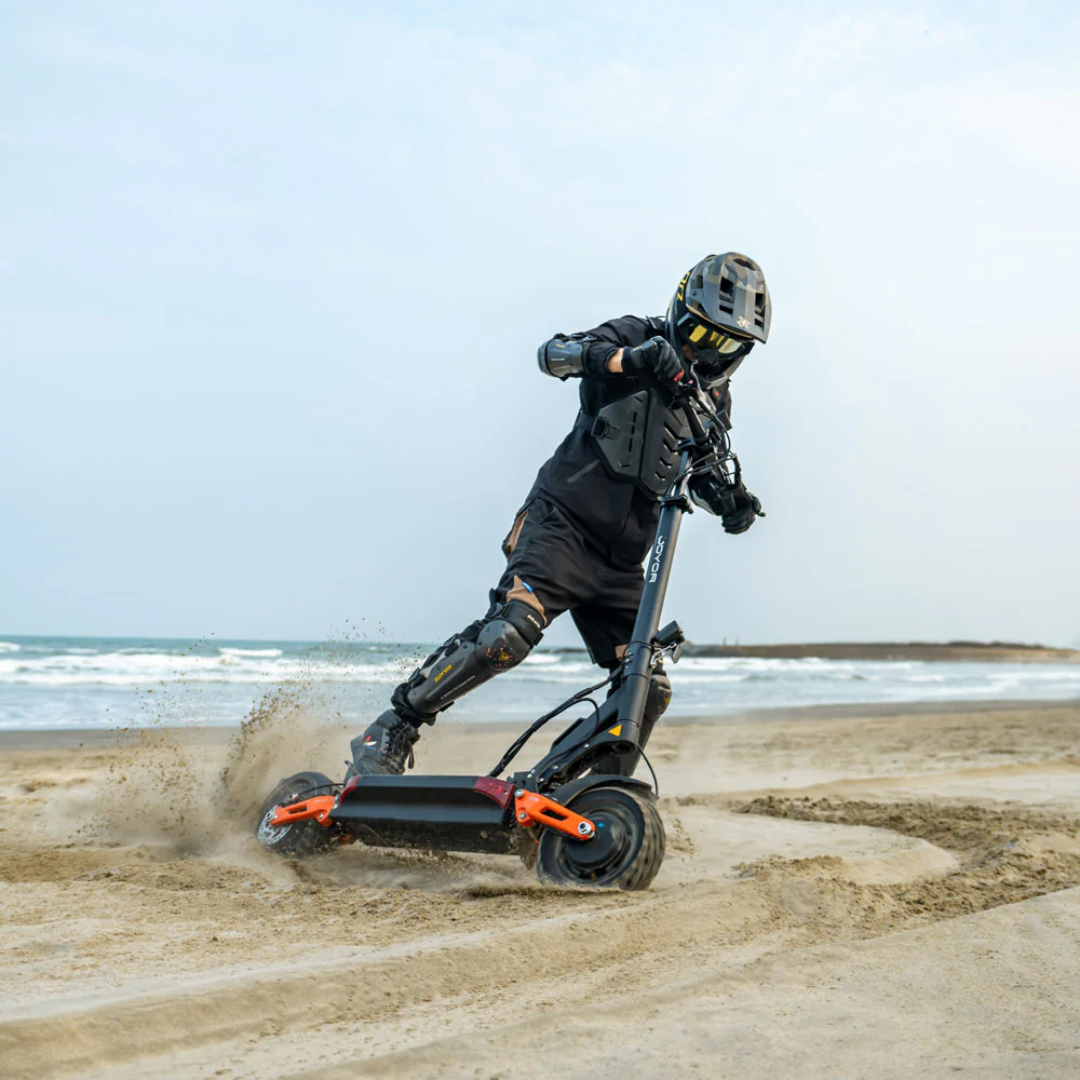 The product in the picture is an electric scooter, model Joyor T10. The picture shows a rider wearing protective gear, riding the scooter on a sandy beach. In the background, the sea and a cloudy sky create a dynamic and adventurous atmosphere.