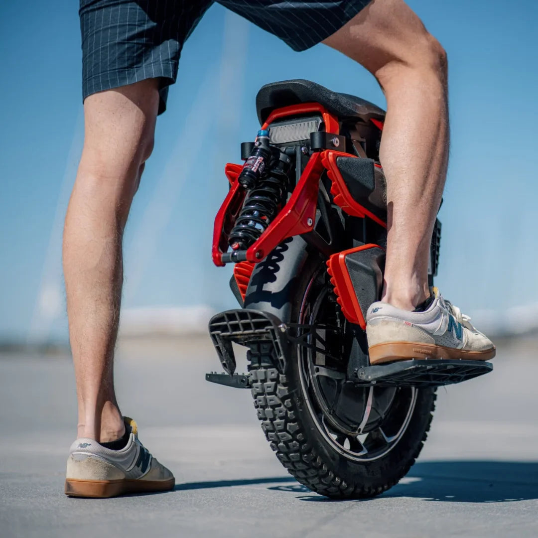 Izdelek je električni monocikel, znamke Kingsong, model S22 Pro. Na sliki je prikazan zadnji del monocikla, ki ima robustno pnevmatiko in vzmetenje. Oseba, ki stoji ob njem, nosi kratke hlače in športne čevlje, kar nakazuje na sproščeno in aktivno uporabo tega prevoznega sredstva. V ozadju je jasno nebo, kar ustvarja prijetno vzdušje.