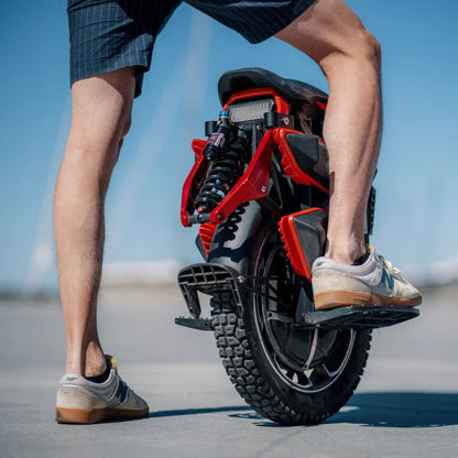 Izdelek je električni monocikel, znamke Kingsong, model S22 Pro. Na sliki je prikazan zadnji del monocikla, ki ima robustno pnevmatiko in vzmetenje. Oseba, ki stoji ob njem, nosi kratke hlače in športne čevlje, kar nakazuje na sproščeno in aktivno uporabo tega prevoznega sredstva. V ozadju je jasno nebo, kar ustvarja prijetno vzdušje.