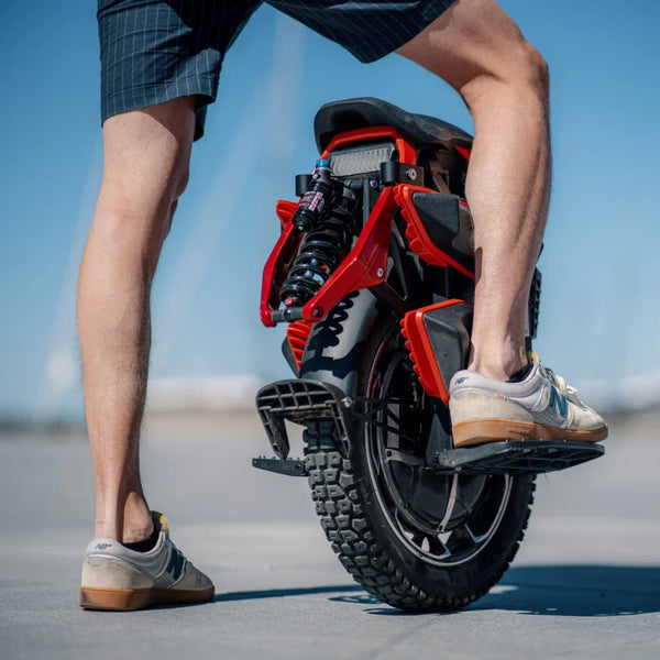 Izdelek je električni monocikel, znamke Kingsong, model S22 Pro. Na sliki je prikazan zadnji del monocikla, ki ima robustno pnevmatiko in vzmetenje. Oseba, ki stoji ob njem, nosi kratke hlače in športne čevlje, kar nakazuje na sproščeno in aktivno uporabo tega prevoznega sredstva. V ozadju je jasno nebo, kar ustvarja prijetno vzdušje.