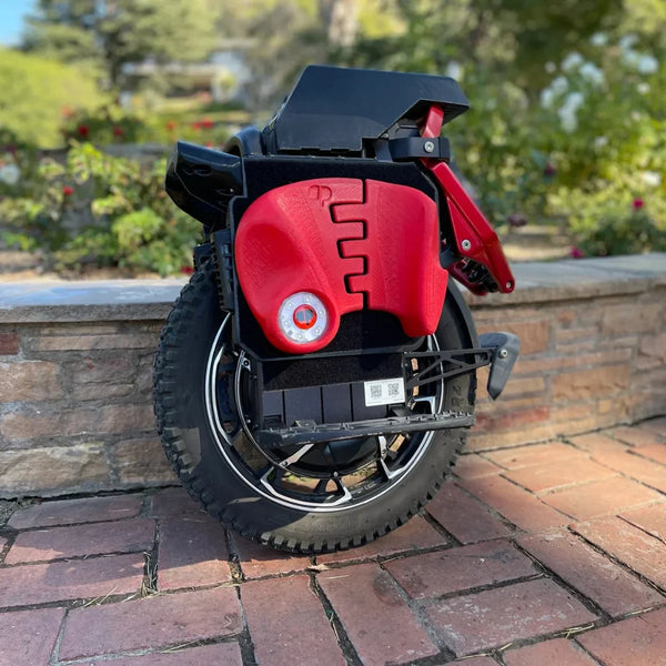 Izdelek na sliki je električni monocikel, model Kingsong S22 Pro. Na sliki je prikazan zložen monocikel, ki ima črno in rdečo barvo. Vidna je velika kolesa z robustnim profilom ter del, kjer se nahajajo kontrole in baterija. Monocikel je postavljen na kamenito površino, v ozadju pa so vidne rastline, kar daje občutek zunanjega okolja.