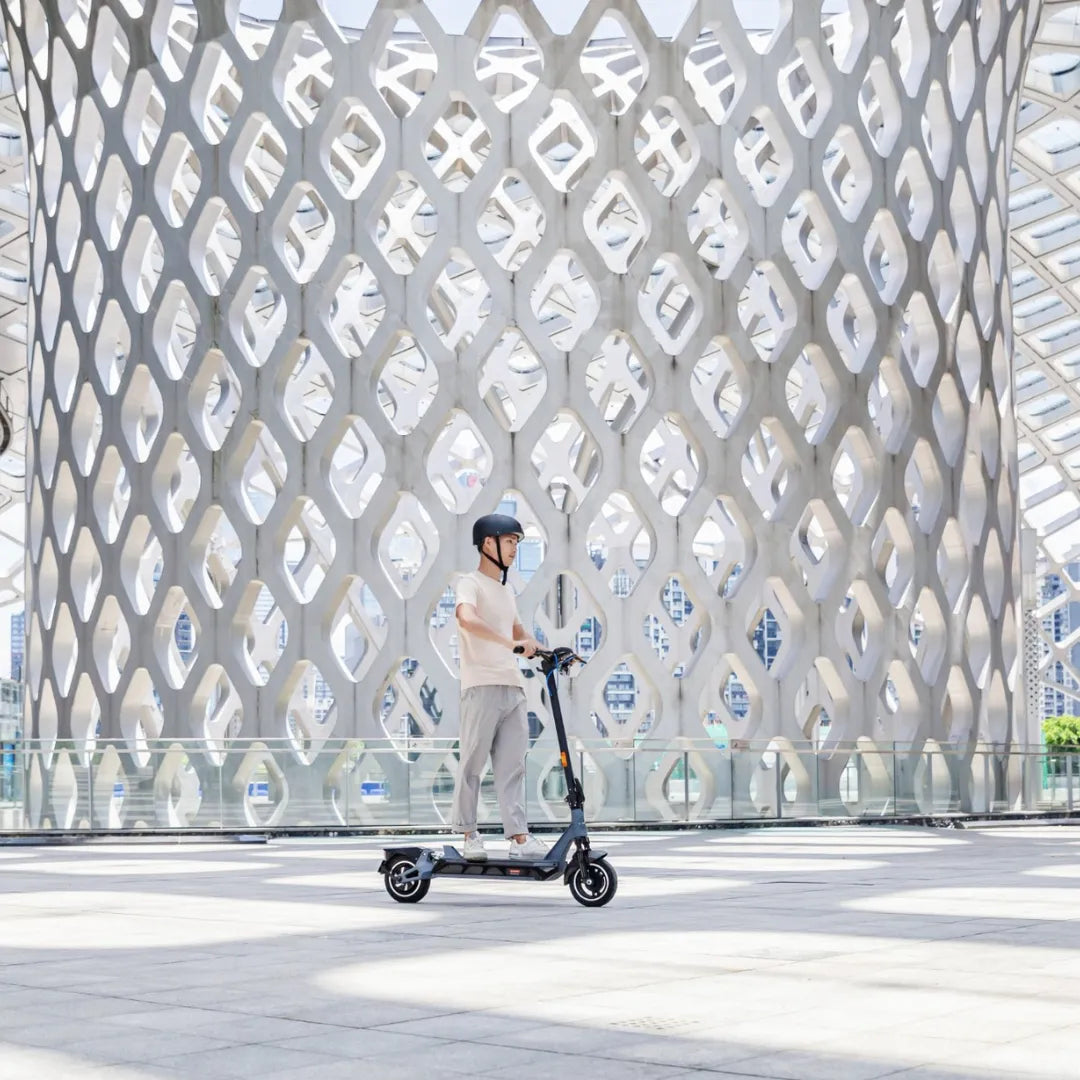 Na sliki je prikazan električni skiro, model Kingsong N12 Pro. Oseba na skiroju nosi čelado in se vozi po modernem, odprtem prostoru z zanimivo arhitekturo v ozadju. Skir je črne barve in ima sodoben dizajn, kar nakazuje na njegovo funkcionalnost in stil.