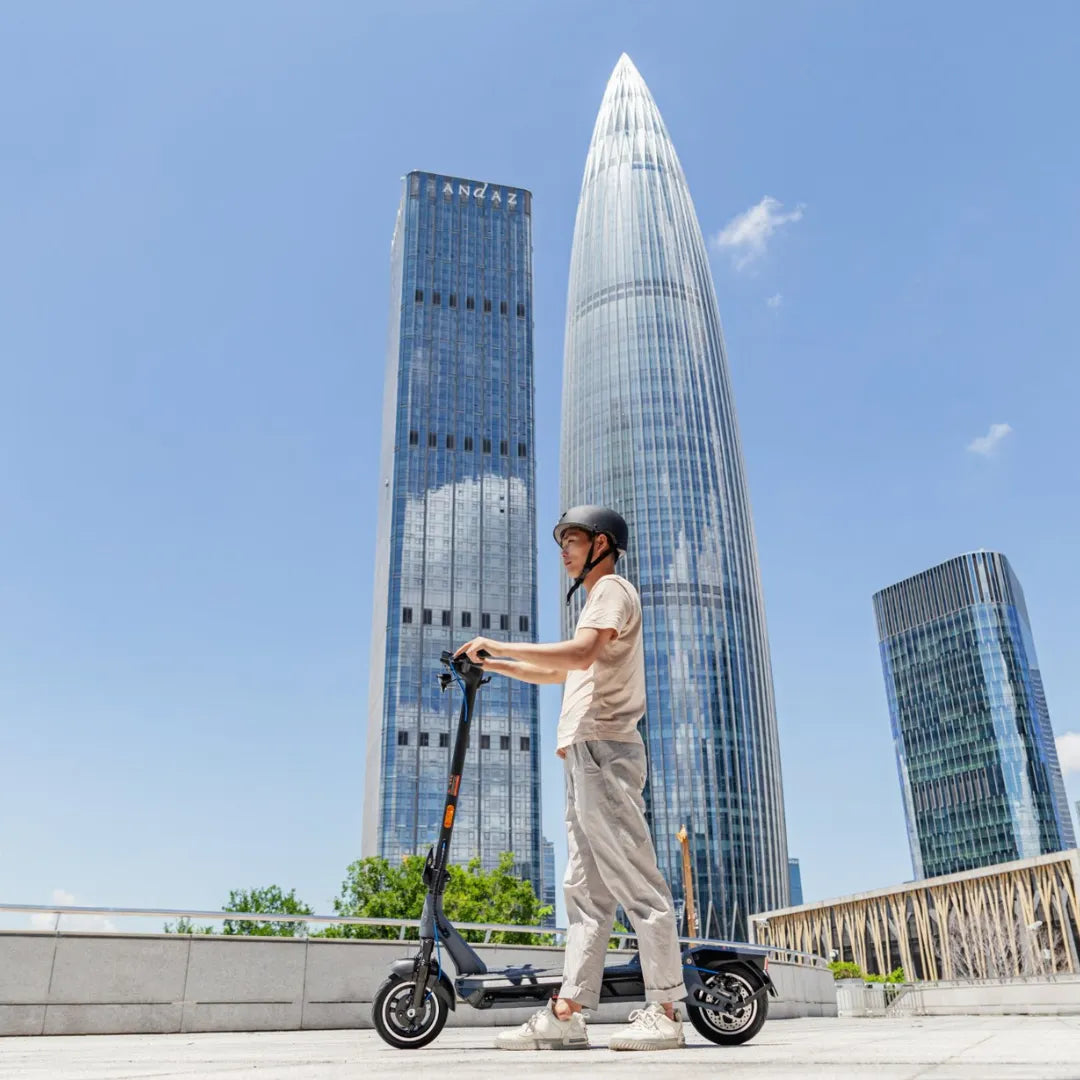 Oseba se vozi z električnim skirojem Kingsong N12 Pro blizu visokih modernih nebotičnikov.