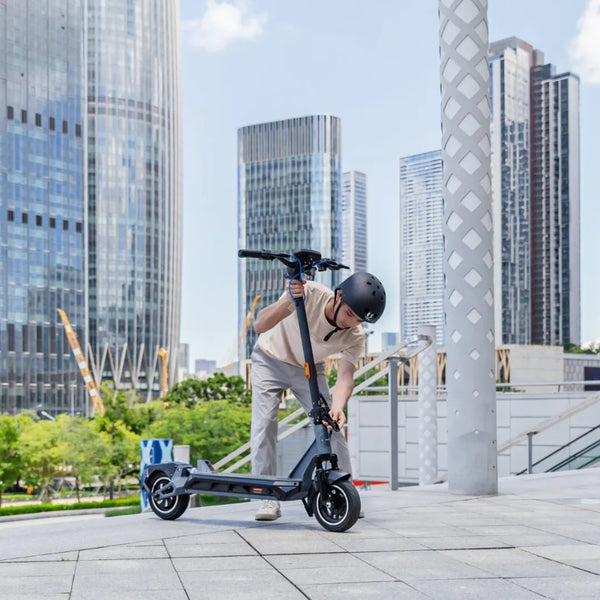 Na sliki je električni skiro, model Kingsong N12 Pro. Vidimo mladega moškega, ki se nagiba nad skirojem, verjetno ga pripravlja za vožnjo. Ozadje prikazuje moderno mestno okolje z visokimi zgradbami in zelenimi površinami. Moški nosi čelado, kar kaže na skrb za varnost med vožnjo.