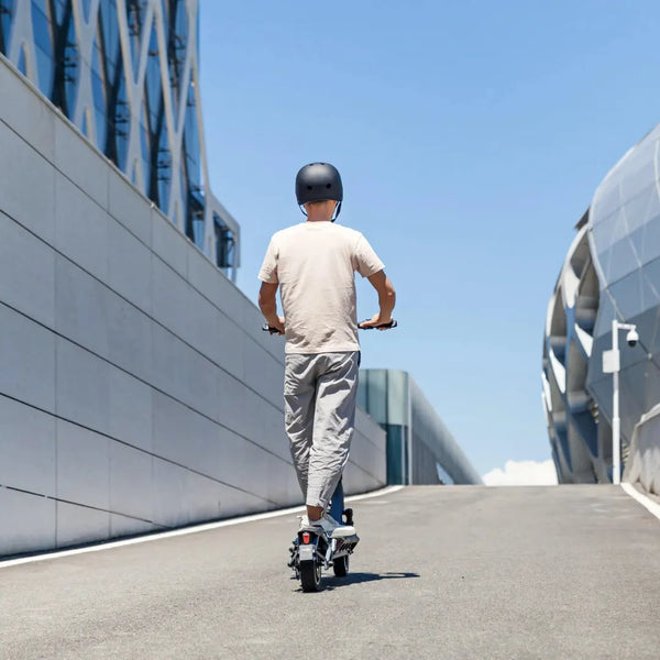 Na sliki je električni skiro znamke Kingsong, model N12 Pro. Oseba vozi skiro po široki cesti, obdanem z modernimi zgradbami in modrim nebom. Opremljena je s čelado, kar nakazuje na zavedanje o varnosti pri vožnji.