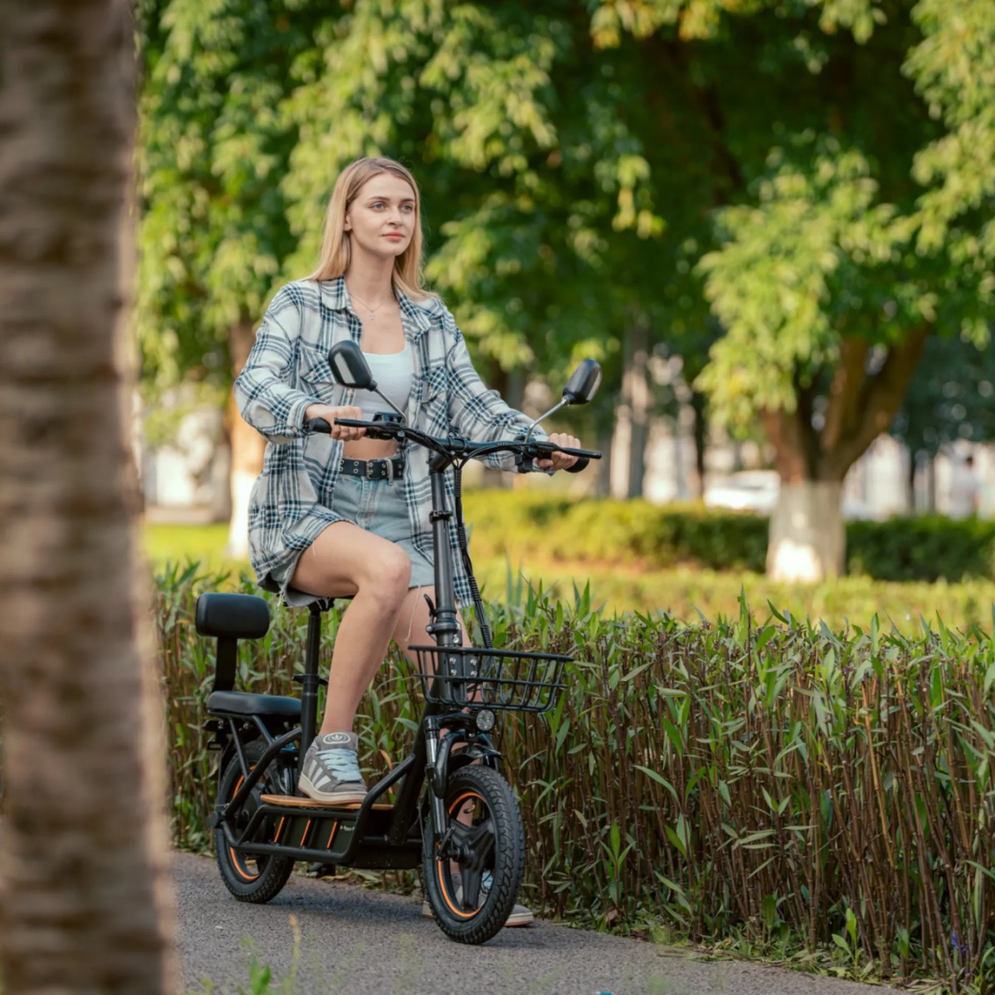 Izdelek na sliki je električni skiro, model Kukirin C1 Pro. Na sliki je vidna mlada ženska, ki sedi na skiroju v parku, obkrožena z zelenjem. Scooter ima nizko zasnovo, široka kolesa in je opremljen z ročaji ter košarico spredaj. Ženska nosi sproščeno obleko in se zdi, da uživa v vožnji.