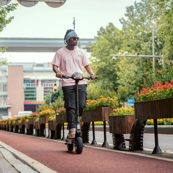 Izdelek na sliki je električni skiro, model Kukirin G2. Na sliki je oseba, ki vozi skiro po urejeni poti ob cvetličnih gredicah. Oseba nosi rožnato majico in sončna očala, okolica pa je zelena in prijetna, kar ustvarja sproščeno vzdušje.