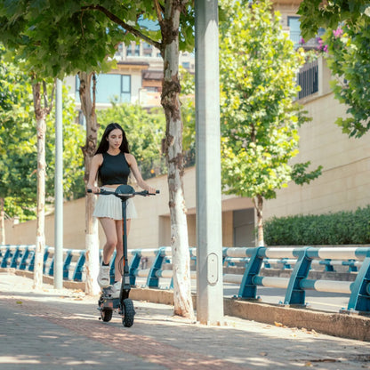 Na sliki je električni skiro znamke Kukirin, model G2. Oseba na skiroju se vozi po urejeni poti ob drevesih, kar kaže na prijetno okolje za vožnjo. Izdelek ima robustne pnevmatike, kar omogoča udobno vožnjo po različnih površinah.