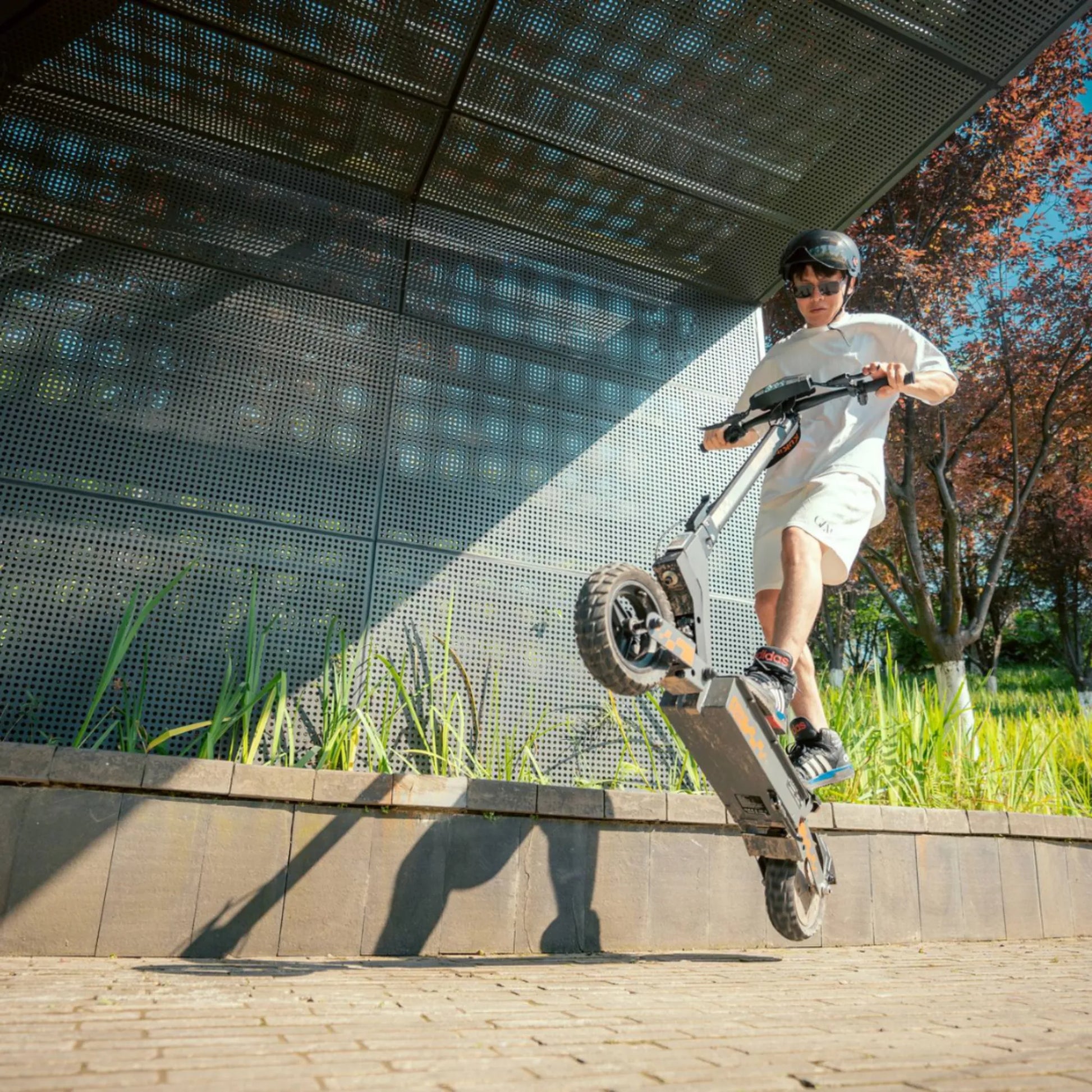 Na sliki je prikazan električni skiro znamke Kukirin, model G2. Uporabnik izvaja akrobacijo, saj skiro dviguje sprednje kolo nad tlakom, medtem ko stoji na pločniku ob zelenju. Opremljen je z robustnimi pnevmatikami, kar nakazuje na primernost za različne terene. V ozadju je modereren arhitekturni element, kar dodaja dinamičnost celotni sliki.