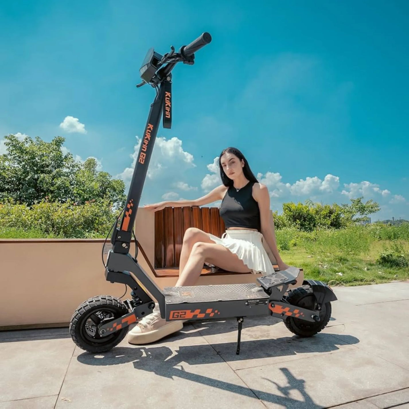 Na sliki je električni skiro znamke Kukirin, model G2. Izdelek ima robustno zasnovo in široke pnevmatike, kar nakazuje na dobro stabilnost in udobje pri vožnji. V ozadju je jasno modro nebo, kar ustvarja prijetno vzdušje, medtem ko se na skiroju nahaja oseba, ki se sproščeno naslanja na ograjo.